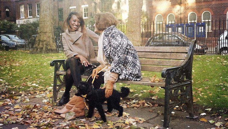 Μια φυσική θέα δύο γυναικών, ντυμένων με ζεστά ρούχα, καθώς συνομιλούν μαζί σε ένα σιδερένιο παγκάκι σε ένα πάρκο του Φθινοπωρινού Λονδίνου. Μία από τις γυναίκες είναι μια ώριμη κυρία που κρατάει απαλά ένα μικρό μαύρο poodle.