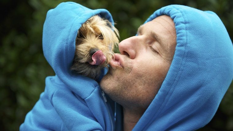 Žmogus bučiuoja savo geriausią draugą šunį, derindamas mėlynus gobtuvus ryškiai žaliame parko fone lauke